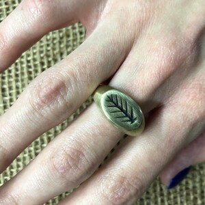 May include: A gold ring with an oval-shaped design featuring a black leaf etched into the surface.