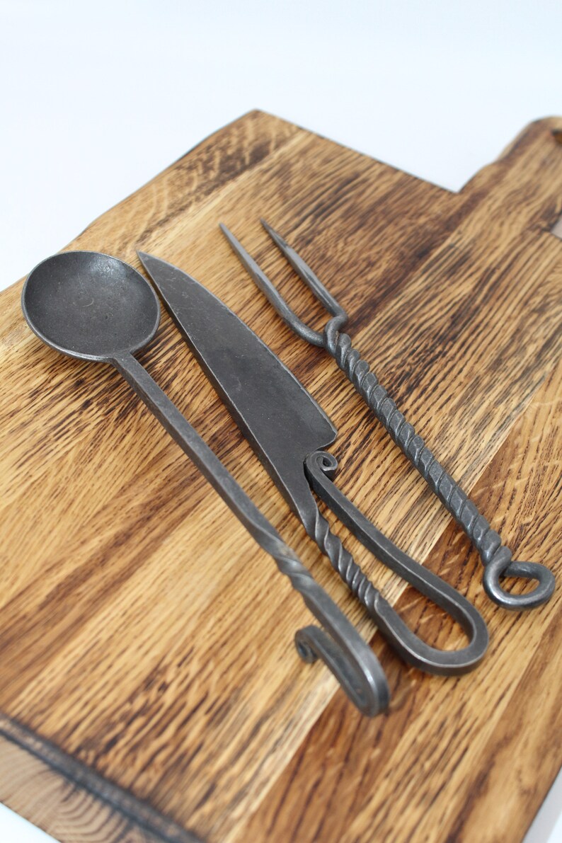 May include: A set of three hand-forged kitchen utensils on a wooden chopping board. The set includes a spoon, a knife, and a fork, all made of black metal.