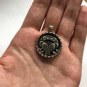 May include: A circular pendant with a design of two birds facing each other. The pendant is made of a gold-colored metal and has a detailed design.