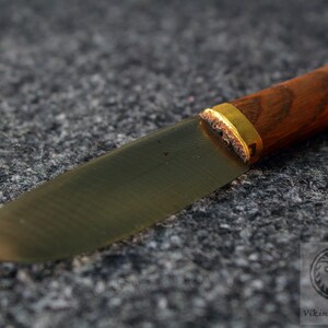 Puede incluir: Primer plano de un cuchillo con mango de madera y una banda de metal dorada. La hoja est&aacute; hecha de metal oscuro y tiene una forma ligeramente curvada.