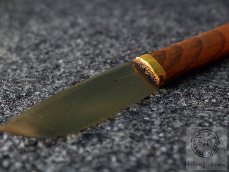 Puede incluir: Primer plano de un cuchillo con mango de madera y una banda de metal dorada. La hoja est&aacute; hecha de metal oscuro y tiene una forma ligeramente curvada.