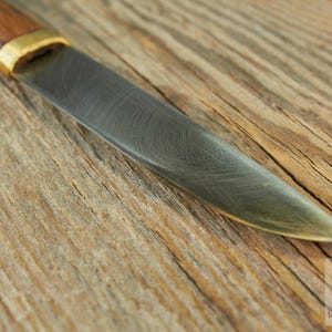 Puede incluir: Primer plano de un cuchillo con mango de madera y hoja de metal. La hoja tiene un acabado martillado y una ligera curva. El cuchillo est&aacute; apoyado sobre una superficie de madera.