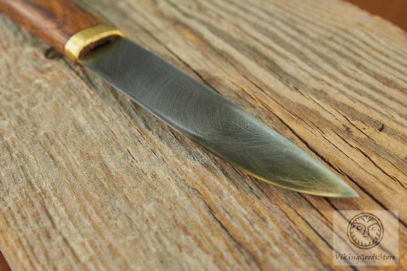Puede incluir: Primer plano de un cuchillo con mango de madera y hoja de metal. La hoja tiene un acabado martillado y una ligera curva. El cuchillo est&aacute; apoyado sobre una superficie de madera.