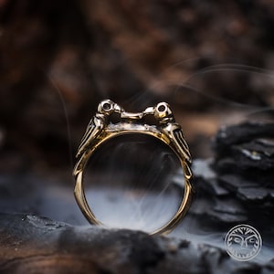 Peut inclure: Une bague en or avec deux oiseaux se faisant face, leurs becs se touchant. Les oiseaux sont détaillés et ont une apparence réaliste. La bague est placée sur un fond sombre avec de la fumée.