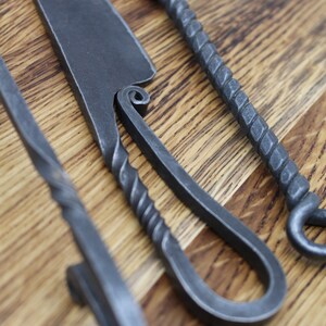 May include: Three hand-forged metal tools with twisted handles. The tools are laid out on a wooden surface. The tools are made of dark grey metal.