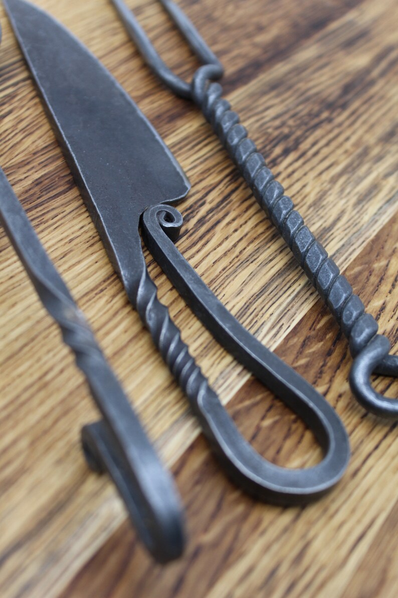 May include: Three hand-forged metal tools with twisted handles. The tools are laid out on a wooden surface. The tools are made of dark grey metal.