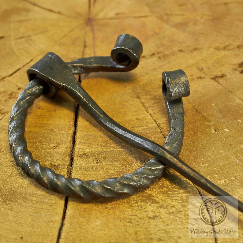May include: A twisted metal Viking brooch with a decorative loop and a long pin. The brooch is made of dark metal and has a rustic finish.