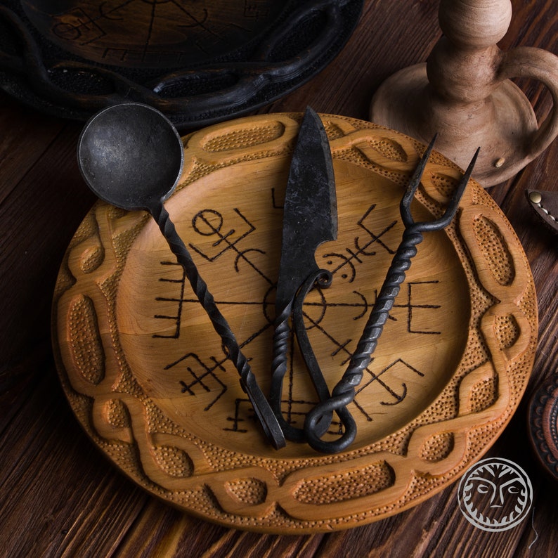 May include: A wooden plate with a carved design of a compass and runes. The plate is decorated with a twisted metal spoon, a knife, and a fork.