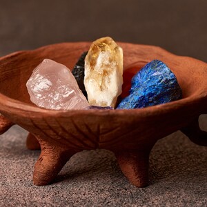 May include: A collection of four crystals, including rose quartz, citrine, amethyst, and lapis lazuli, are displayed in a small, brown, three-legged ceramic bowl.