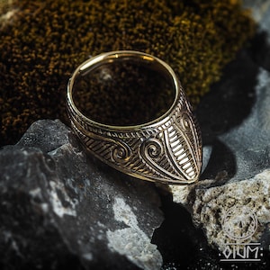 Puede incluir: Un anillo de oro con un diseño detallado e intrincado. El anillo está hecho de una banda gruesa con una parte superior puntiaguda y triangular. El diseño presenta una serie de patrones y líneas en espiral.