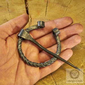 May include: A twisted metal ring with a long, pointed pin. The ring is made of dark metal and has a textured surface. The pin is also made of dark metal and has a smooth surface.