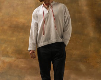 May include: A light gray long-sleeve shirt with a red drawstring neckline and black pants. The shirt has a relaxed fit. The model is barefoot. The background is a mottled brown.