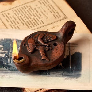 May include: A brown ceramic oil lamp with a handle and a detailed relief of a figure. The lamp is lit, with a bright yellow flame. The lamp is resting on a vintage postcard and old paper.