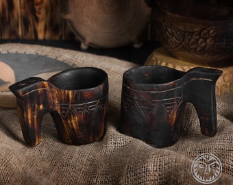May include: Two dark brown wooden drinking cups with carved designs. The cups have a handle that resembles a stylized animal head. The cups are sitting on a burlap surface.