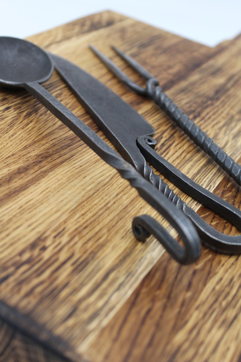 May include: A set of three hand-forged kitchen utensils on a wooden chopping board. The set includes a spoon, a knife, and a fork.