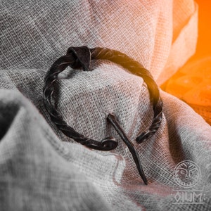 May include: A close-up of a black metal Viking-style brooch with a twisted wire design. The brooch is lying on a piece of white fabric.