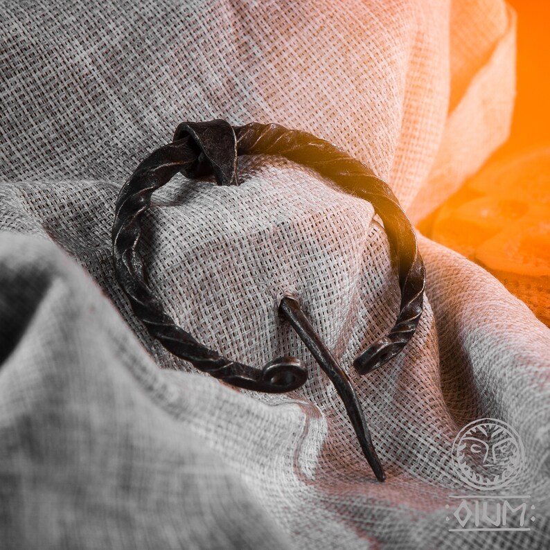 May include: A close-up of a black metal Viking-style brooch with a twisted wire design. The brooch is lying on a piece of white fabric.
