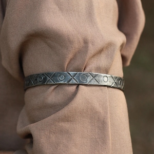 Pode incluir: Uma pulseira de metal prateado com um padrão gravado de círculos e formas de X alternados. A pulseira é usada sobre um fundo de tecido bege, sugerindo um tema rústico ou histórico. O design é simétrico e detalhado.