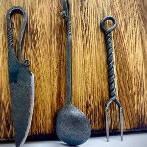 May include: A set of three hand-forged kitchen utensils: a knife, a spoon, and a fork. The utensils are made of dark metal and have a twisted design. They are displayed on a wooden surface.