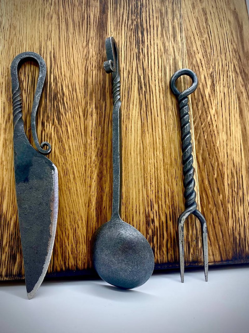 May include: A set of three hand-forged kitchen utensils: a knife, a spoon, and a fork. The utensils are made of dark metal and have a twisted design. They are displayed on a wooden surface.