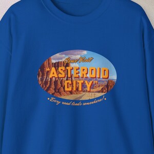 May include: A royal blue sweatshirt with a vintage-style graphic print of a desert landscape. The text on the graphic reads "Come Visit 'Asteroid City' - Every road leads somewhere!"