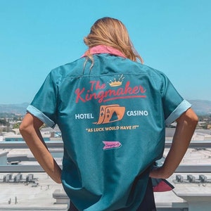 May include: A teal blue bowling shirt with a retro design featuring the text "The Kingmaker Hotel Casino" and a graphic of playing cards. The text "As luck would have it" is printed below the graphic.