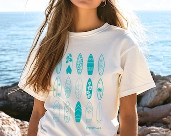 Puede incluir: Camiseta color crema con un diseño de tabla de surf turquesa. El gráfico incluye varias ilustraciones de tablas de surf con diferentes patrones y formas. El texto "Dragonfly Surf Co." está impreso en la parte inferior del diseño. La camiseta es de material suave.