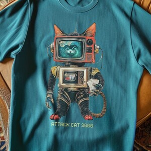 May include: Teal blue t-shirt with a graphic print of a cat wearing a television for a head and holding a cup. The text "Attack Cat 3000" is printed below the graphic.