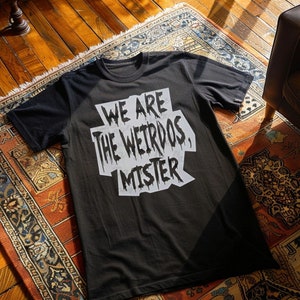 Peut inclure: T-shirt noir avec l'inscription blanche "WE ARE THE WEIRDOS, MISTER" dans une police de caractères vieillie. Le t-shirt est posé sur un tapis à motifs aux tons rouges, oranges et bruns. Col rond.