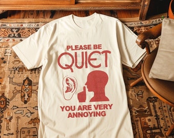 May include: Cream-coloured t-shirt with the words "PLEASE BE QUIET" in red, and a graphic of an ear, a person shushing, and the text "YOU ARE VERY ANNOYING". The shirt is laid flat on a patterned rug.