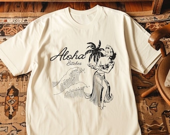 May include: A white t-shirt with a black graphic print of a woman in a hula skirt, palm trees, and waves. The text "Aloha Bitches" is printed on the shirt.