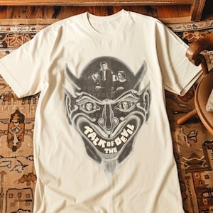 May include: A white t-shirt with a graphic print of a devil's face. The face is made up of a group of people in a photo. The text "Talk of the Devil" is printed below the face.