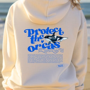 May include: A cream-coloured hoodie with a blue and white graphic of a killer whale. The graphic says "Protect the Orcas" with a heart symbol. The text below the graphic says "End Orca Captivity".