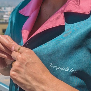 May include: A teal blue and pink button-up shirt with the text "Dangerfolk Inc." printed on the front. The shirt has a relaxed fit and a collar.
