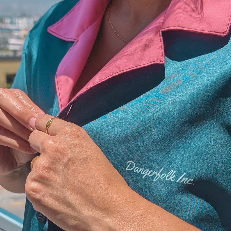 May include: A teal blue and pink button-up shirt with the text "Dangerfolk Inc." printed on the front. The shirt has a relaxed fit and a collar.