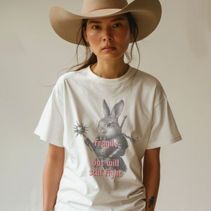 K&ouml;nnte beinhalten: Wei&szlig;es T-Shirt mit einer detaillierten Illustration eines Kaninchens in R&uuml;stung, das eine Keule und ein Schwert h&auml;lt. Der Text "fragile but will still fight" ist in Rosa gedruckt. Die Person tr&auml;gt einen beigen Cowboyhut.