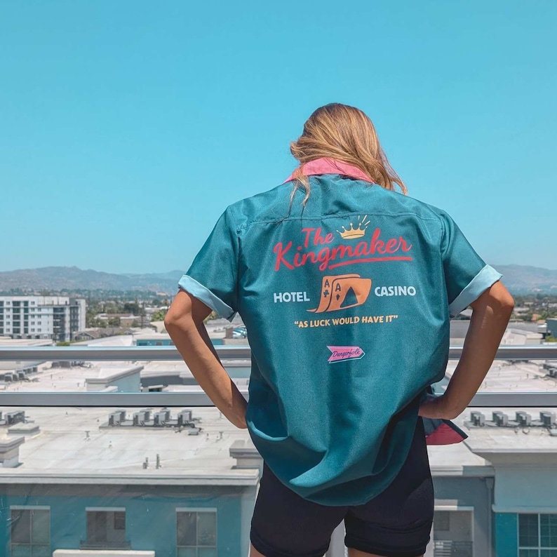 May include: A teal blue bowling shirt with a retro design featuring the text "The Kingmaker Hotel Casino" and a graphic of playing cards. The text "As luck would have it" is printed below the graphic.