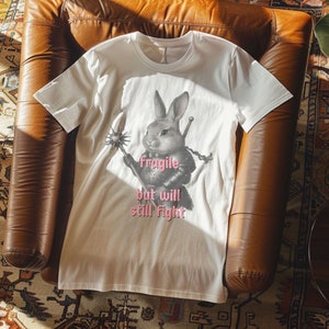 K&ouml;nnte beinhalten: Cremefarbenes T-Shirt mit einer detaillierten Illustration eines Kaninchens in R&uuml;stung, das eine Waffe und ein Schwert h&auml;lt. Der Text "fragile but will still fight" ist rosa unter dem Kaninchen aufgedruckt. Das Shirt ist aus weichem Material.