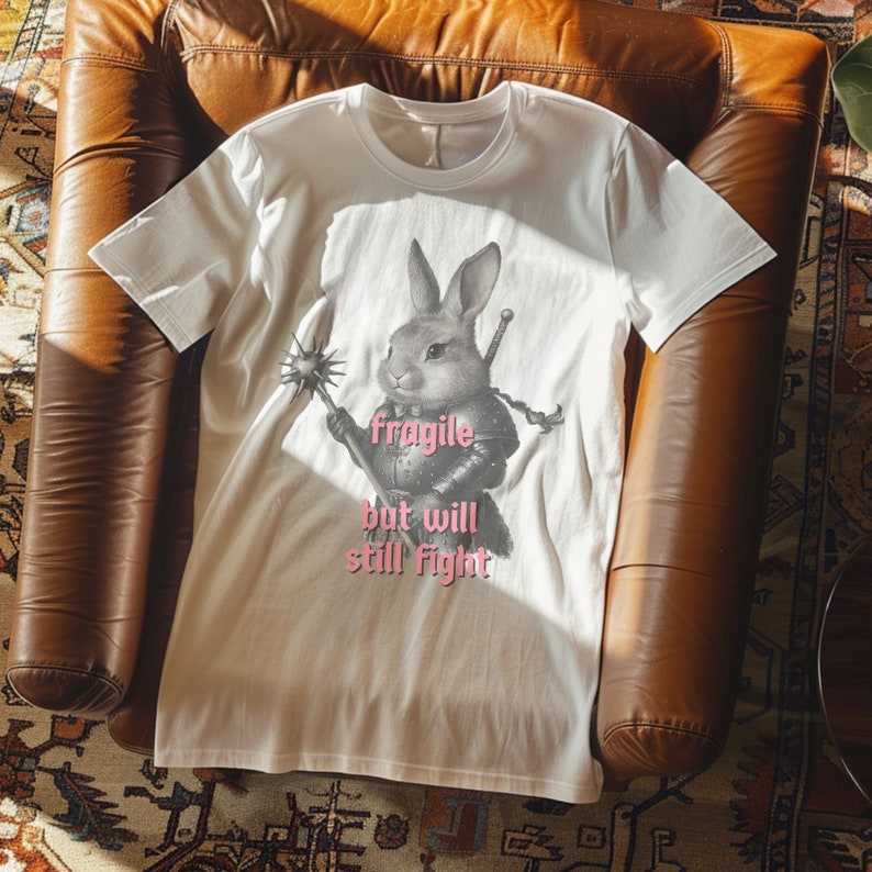 K&ouml;nnte beinhalten: Cremefarbenes T-Shirt mit einer detaillierten Illustration eines Kaninchens in R&uuml;stung, das eine Waffe und ein Schwert h&auml;lt. Der Text "fragile but will still fight" ist rosa unter dem Kaninchen aufgedruckt. Das Shirt ist aus weichem Material.