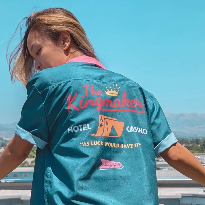 May include: A teal blue bowling shirt with a pink collar and a graphic design on the back. The graphic design features the text "The Kingmaker", a crown, playing cards, and the text "Hotel", "Casino", and "As luck would have it".