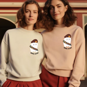May include: Two women wearing light pink and light green sweatshirts with a cartoon tooth wearing a scout uniform and the text "Ghoul Scout".