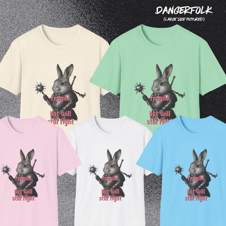 K&ouml;nnte beinhalten: Verschiedene T-Shirts in Creme, Mintgr&uuml;n, Rosa, Wei&szlig; und Hellblau. Jedes Shirt zeigt eine Grafik eines Kaninchenkriegers mit dem Text "Fragile but will still fight". Der Markenname "DANGERFOLK" ist sichtbar.