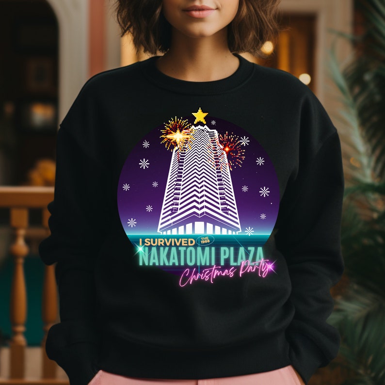 May include: Black sweatshirt featuring a graphic of a skyscraper decorated with a star and fireworks, with the text "I Survived Nakatomi Plaza Christmas Party". The design is set within a purple and blue circle.