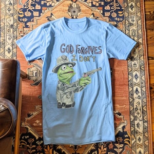 May include: Light blue t-shirt with a cartoon image of a frog wearing a police hat and holding a gun. The text "God Forgives I Don't" is printed on the shirt.