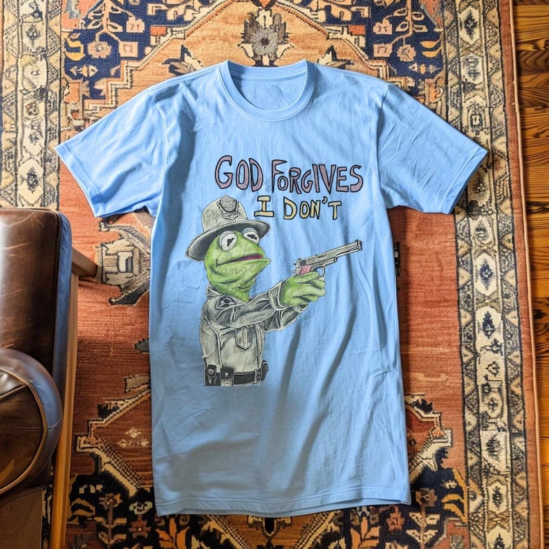 May include: Light blue t-shirt with a cartoon image of a frog wearing a police hat and holding a gun. The text "God Forgives I Don't" is printed on the shirt.