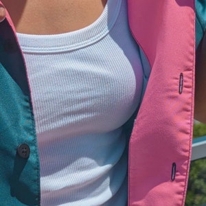 May include: A white ribbed tank top layered under a teal and pink button-up shirt. The shirt has a contrasting pink panel on the right side.