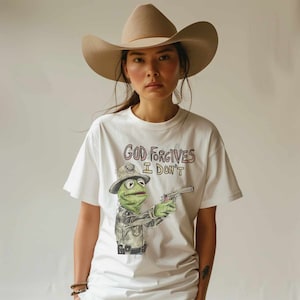 May include: A white t-shirt with a green frog wearing a police hat and holding a gun. The shirt has the text "God Forgives I Don't" printed on it.