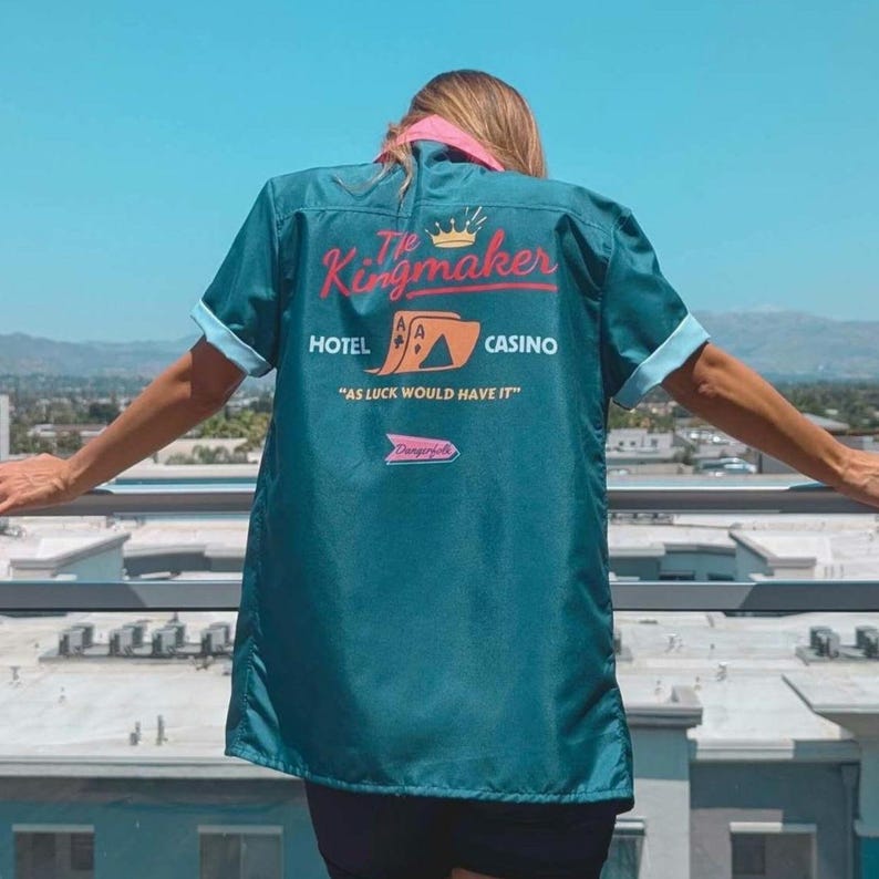 May include: A teal blue bowling shirt with a pink collar and a graphic print on the back. The graphic features the text "The Kingmaker", a crown, playing cards, and the words "Hotel", "Casino", and "As luck would have it".