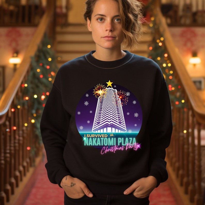 May include: Black crew neck sweatshirt with a graphic featuring a tall building, fireworks, and the text "I Survived Nakatomi Plaza Christmas Party". The design is set within a purple and blue circle with snowflakes.