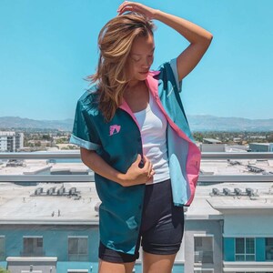 May include: A woman wearing a teal and light blue button-up shirt with a pink collar and a white tank top underneath. She is wearing black shorts and has her hair pulled back.